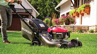 A person in jeans pushes a lawn mower in a residential front yard.  