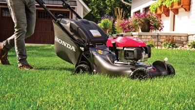A person in jeans pushes a lawn mower in a residential front yard.  